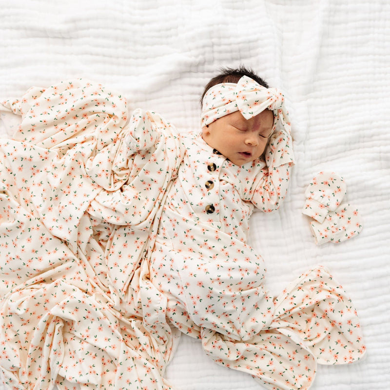 lottie headband bundle newborn girl wearing knotted gown, swaddle blanket, knot headband and mittens laying on bed.
